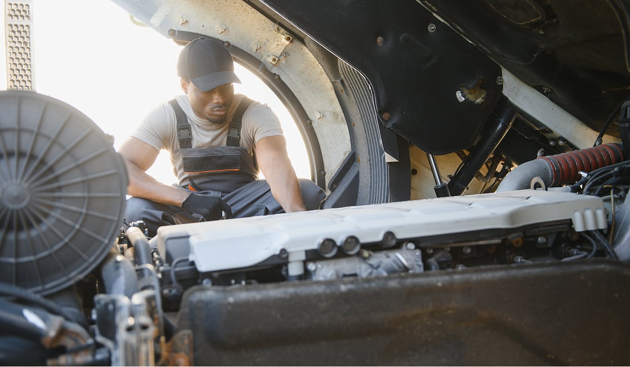 Mechanic Wearing a Cap and Overalls Crouching in the Open Engine Bay of a Large Vehicle Inspecting the Engine Components