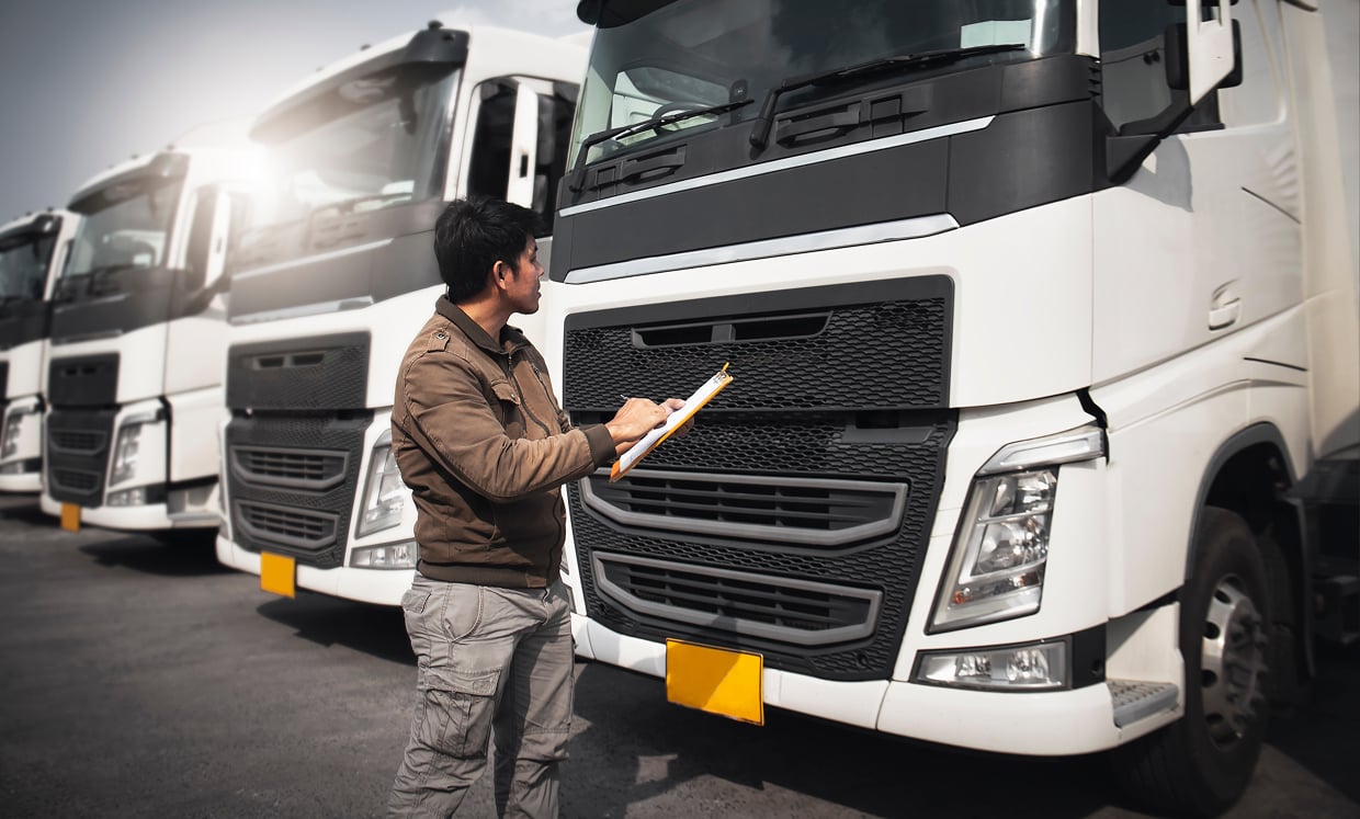 Man in a Brown Jacket with a Clipboard Inspecting a Row of White Semi trucks in a Parking Lot Piece by Piece for Fleet Maintenance Inspection