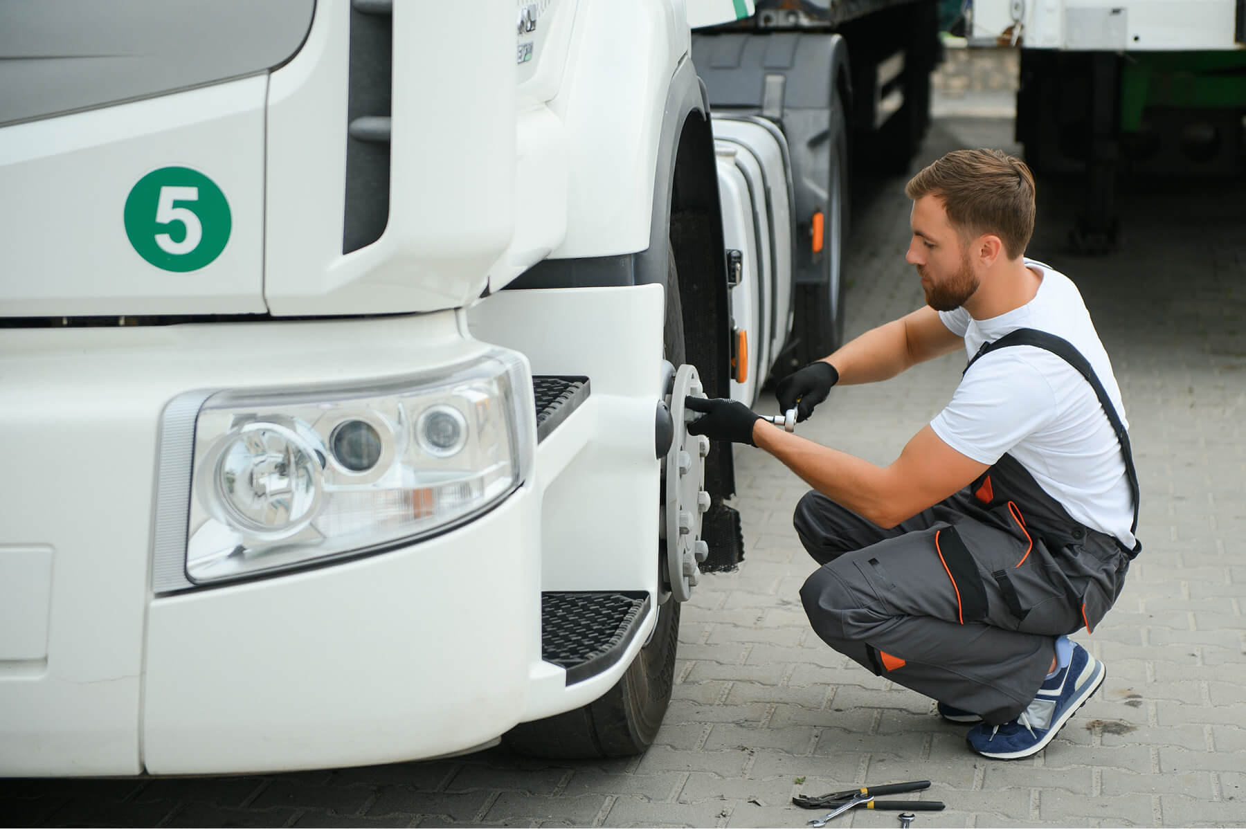 Male Mechanic Kneels by a White Truck Tightening the Front Wheel with a Wrench in His Gloved Hands