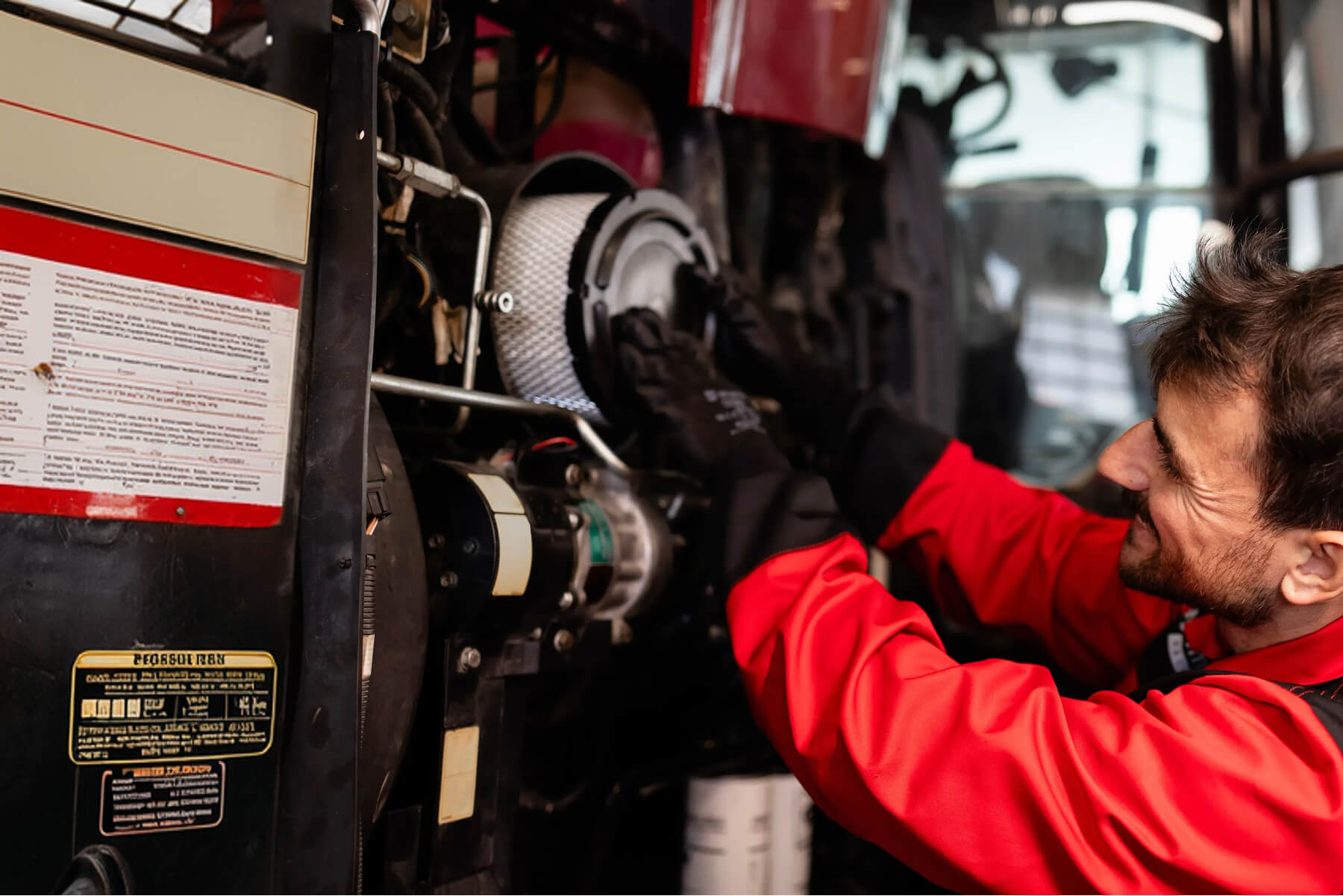 Mechanic in Red Coveralls Inspecting a Bus Engine Kneeling Beside Exposed Components and Labels in a Workshop