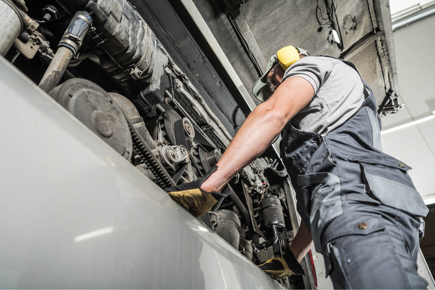 Maintenance Worker in Gray Overalls and Yellow Ear Protection Inspects a Complex Engine Inside a Large Machine