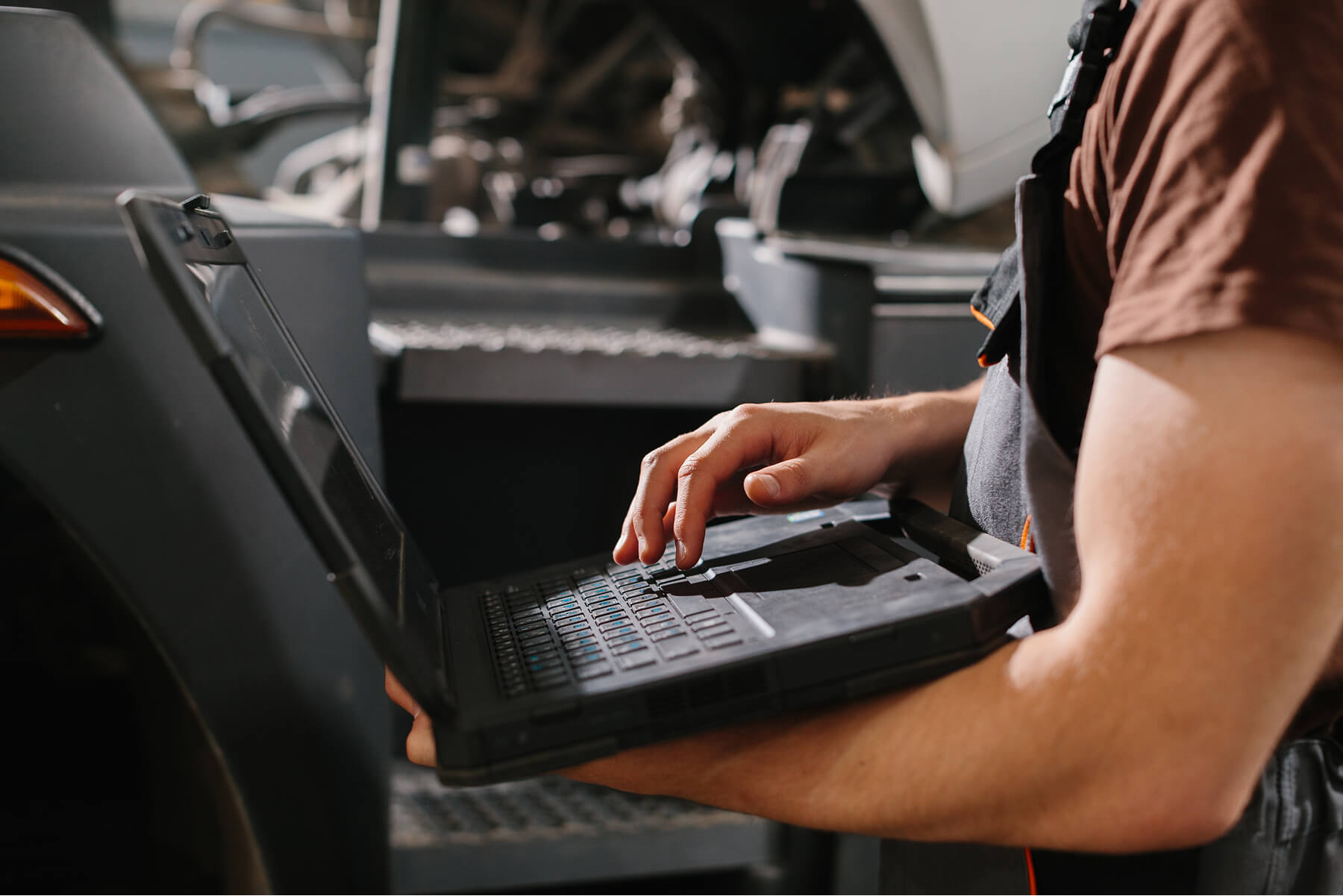 Auto Technician in a Brown Shirt Typing on a Rugged Laptop Beside an Open car Hood in a Workshop
