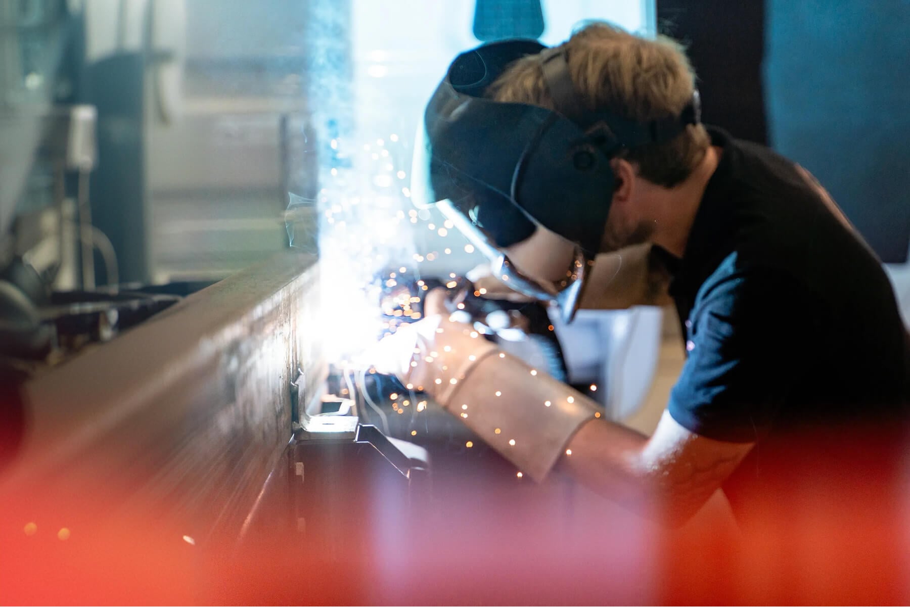 Welder in Protective Helmet and Gloves Bending over Metal Sparks Flying As They Weld a Long Seam in a Workshop