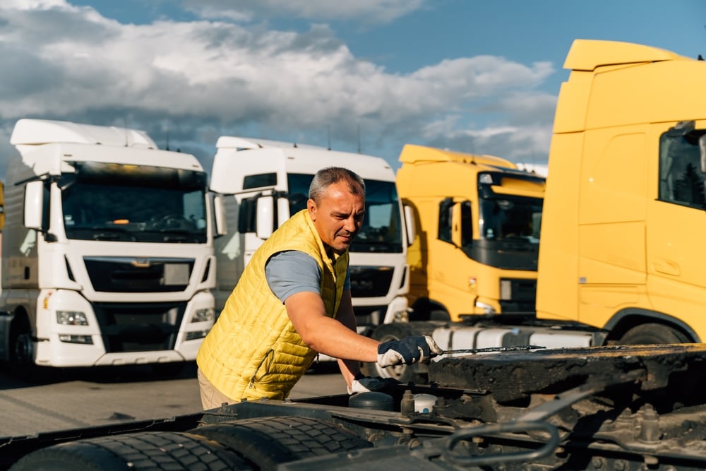 Maintenance worker in a yellow vest and gloves tightening a truck engine in a yard with white and yellow trucks ahead of him.