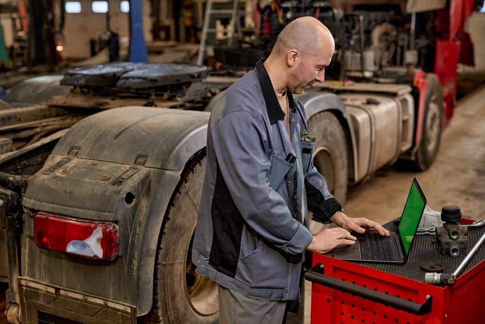 Mechanic in a gray uniform typing on a laptop at a red tool cart beside a dusty semi-truck in a workshop.