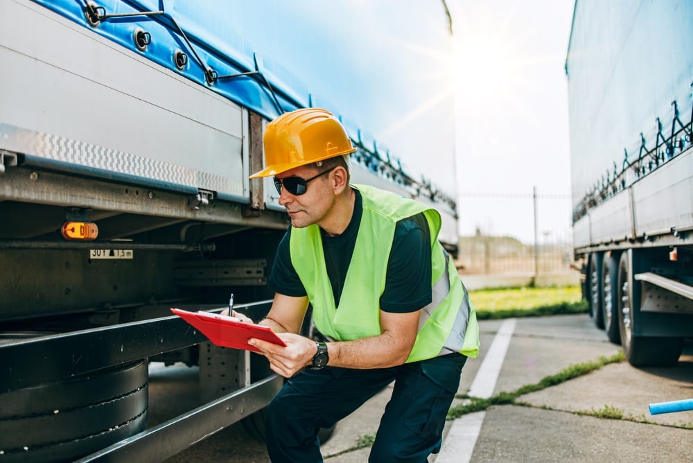 Construction worker in a high-visibility vest and orange hard hat crouches beside a trailer, writing on a red clipboard under bright sun.