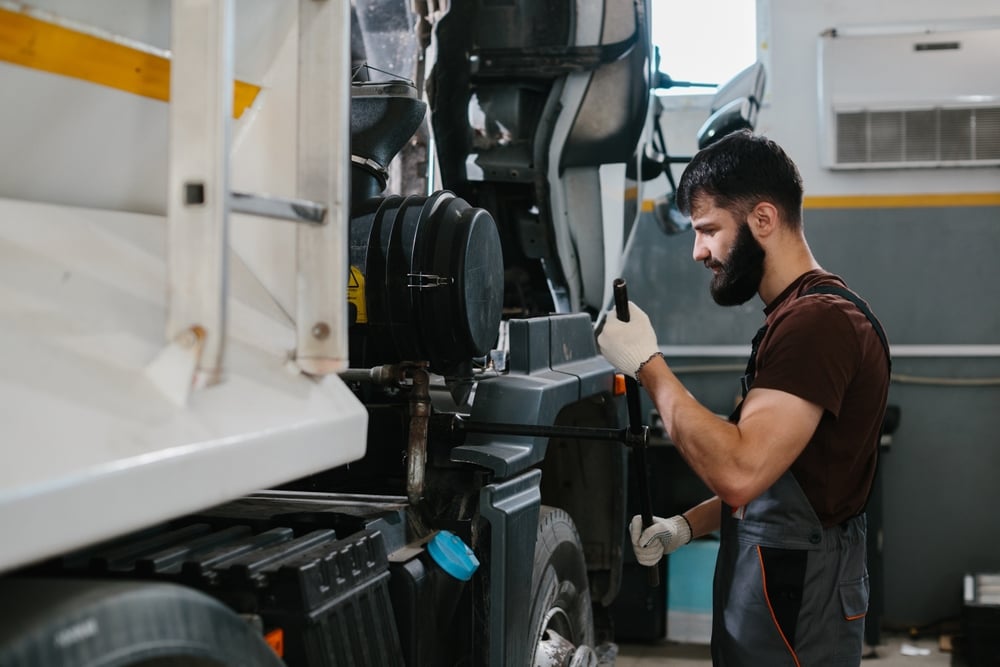 Male mechanic with a beard in brown shirt and gray overalls inspecting a heavy truck engine in a workshop.