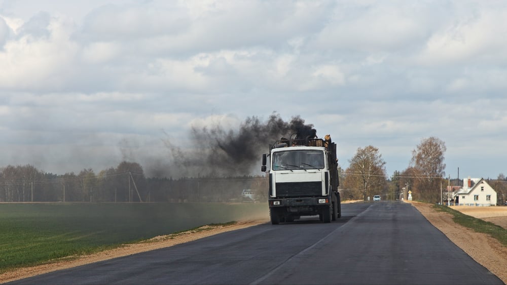 White flatbed truck on a rural road emitting thick black exhaust.