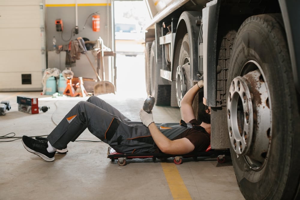 Mechanic lying on a mechanic’s creeper beneath a large truck, inspecting with a flashlight in a workshop.
